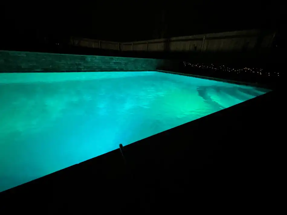 A pool lit teal at night beside a stacked stone retaining wall and string lights, an Oklahoma pool Cardinal Pools services
