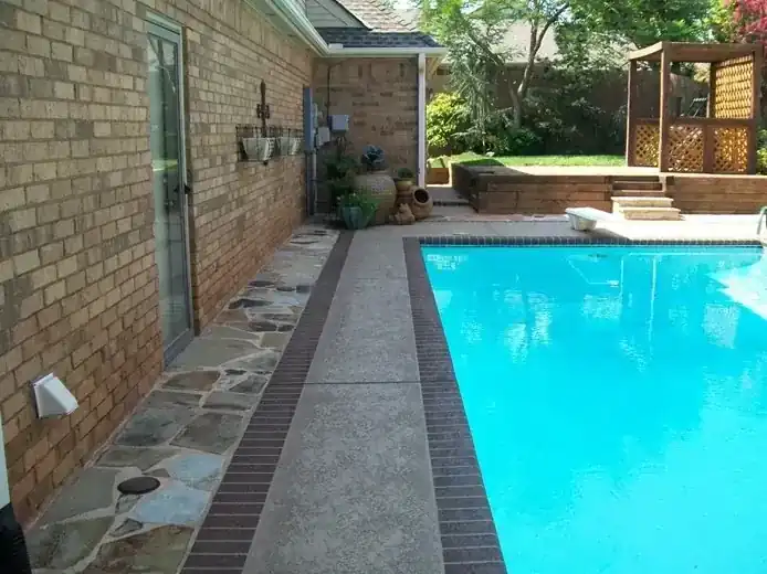 A narrow side yard rectangular pool with a flagstone path and banded Sundek coping beside a brick home